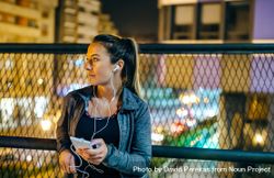 Female athlete with earphones looking away while resting over bridge after training at night in town 5zrR6m