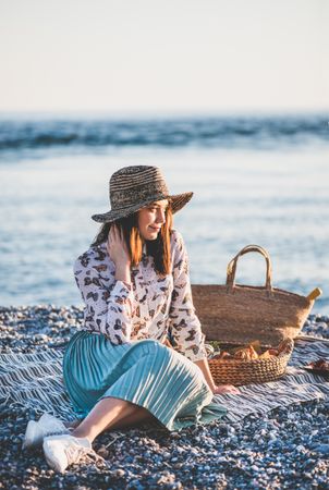 Young woman in hat sitting by the ocean with a picnic