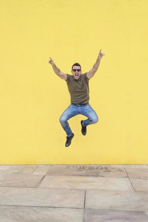 Excited male jumping outside in front of yellow wall with arms up