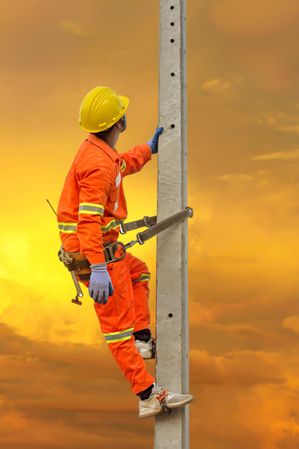 Back view of man in overall and bump cap climbing a pole at sunset