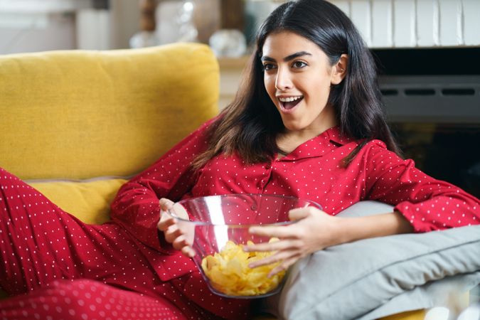 Woman in red pajamas lying on sofa and eating chips