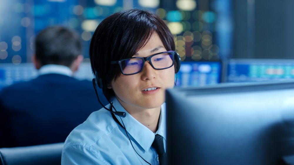 In the Network Operations Center Senior Trader Teaches Young Associate to Makes Personal Client Call with a Headset.