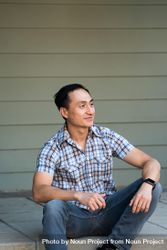 Man in plaid shirt sitting on steps in front of house smiling and looking away bE9qnb