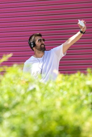 Latino man in headphones taking selfie with phone in front of pink wall