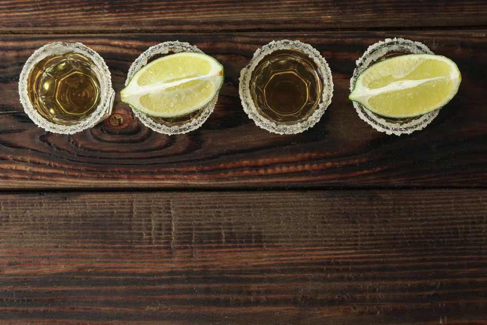 Tequila shots with salt and lime slices on wood table, top view
