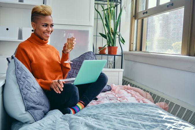 Happy Black woman smiling at a laptop with champagne flute in light filled living room