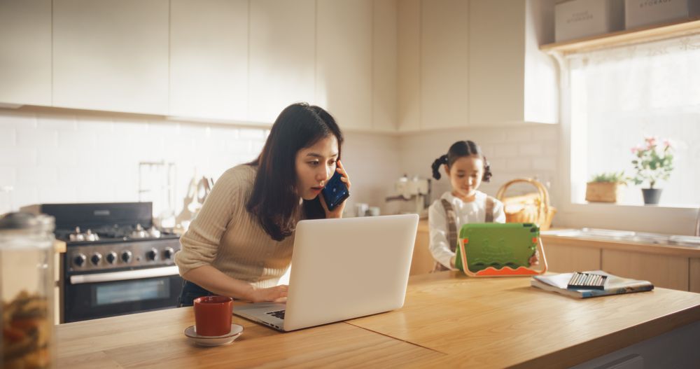 Portrait of Korean Mother Using Laptop and Smartphone To Work from Home While her Little Daughter is Using Digital Tablet.