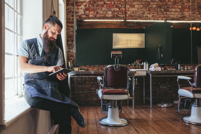 Barber with digital tablet sitting on window sill of barbershop