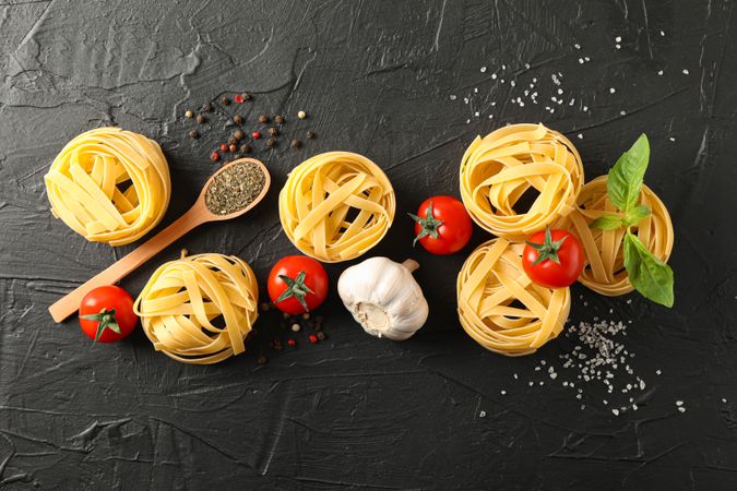 Flat lay composition with pasta, tomatoes, salt, garlic and spices on dark background, space for text
