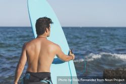 Male surfer holding blue board standing in front of the ocean 43kYP4