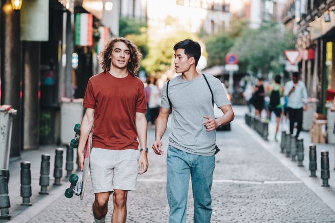 Cool multiracial male friends chatting while walking on street with longboard