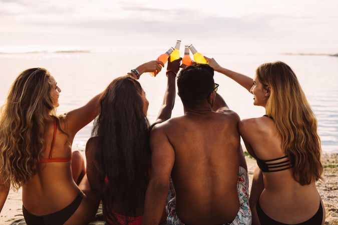 Rear view of group of friends sitting together on beach toasting bottles of drinks