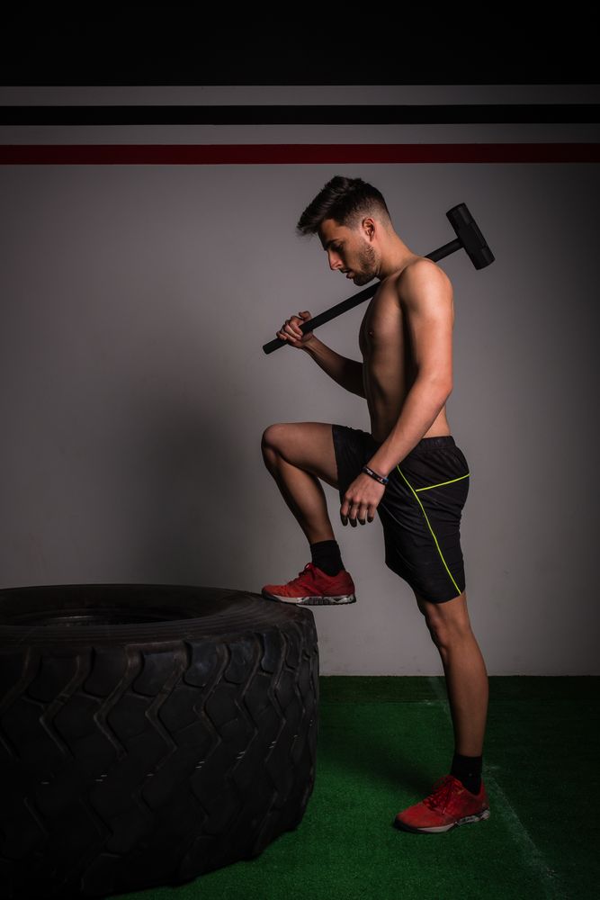 Shirtless guy concentrated in sportswear with hammer hitting on big tire in gym