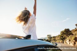 Rearview of woman standing out of sunroof on sunny day and celebrating 0vaWpb