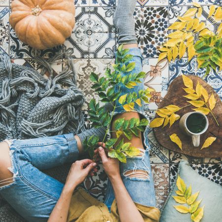 Woman sitting on colorfully tiled balcony with fall leaves, squash, and mug, square crop