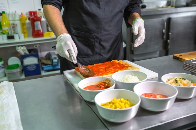Anonymous male chef with gloves spreading tomato sauce over pizza in commercial kitchen