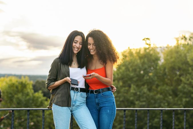 two young women smiling and using her mobiles on a terrace. with the sunset