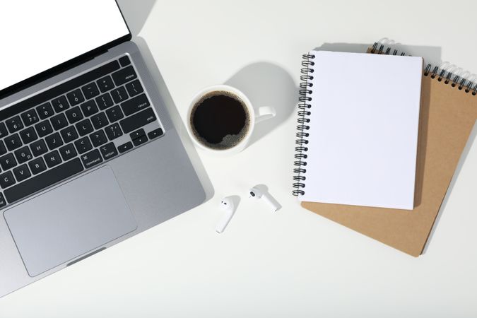 Top view of table with open laptop with mockup screen, earbuds, coffee, and blank notebooks