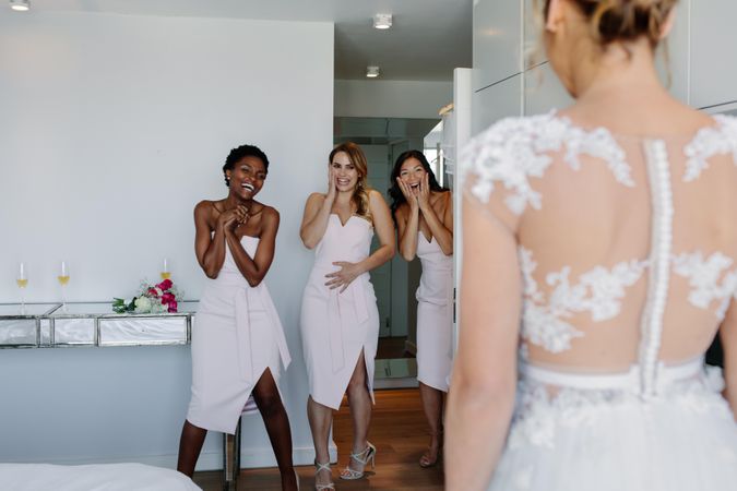 Surprised bridesmaids looking at beautiful bride in wedding gown in hotel room