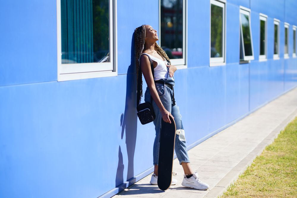 Female in denim overalls standing with skateboard while leaning on blue wall