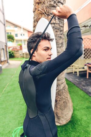 Young surfer man with surfboard closing zipper on wetsuit