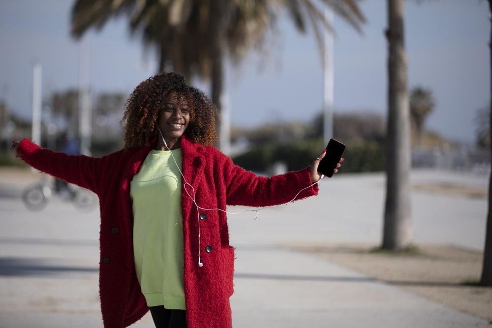 Happy female in colorful clothes dancing to music on phone on street with palm trees
