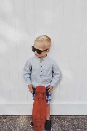 Young blond boy leaning against a wall with red skateboard