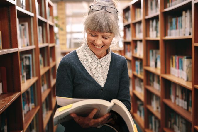 Cheerful mature woman standing in a library looking at a textbook