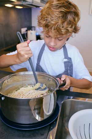 Focused boy stirring cooked spaghetti in saucepan on stove