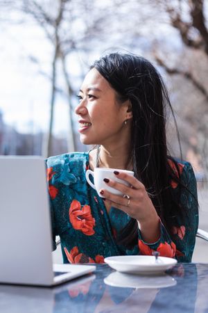 Long-haired brunette Asian woman having a coffee on a terrace of a cafe while is working with a laptop