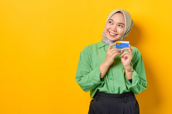 Happy Muslim woman in headscarf and green blouse holding credit card with both hands looking away