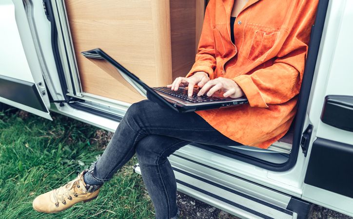 Female in orange coat sitting on step of van with laptop