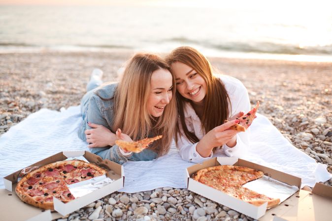 Two smiling women eating pizza on seashore
