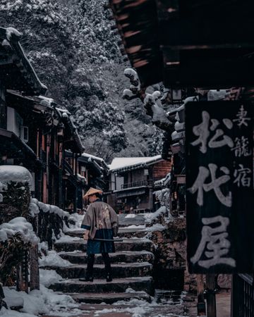 Back view of Japanese person with conical hat walking in snow-covered ancient alley in Japan