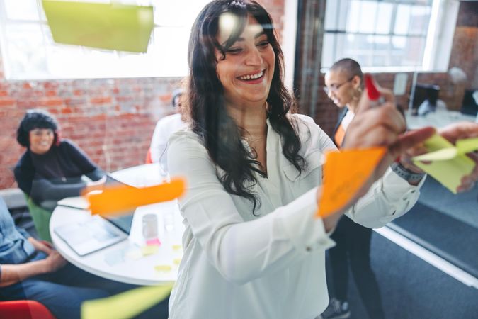 Smiling businesswoman sticking adhesive notes to a glass wall with her colleagues in the background