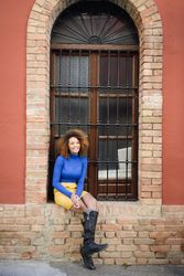 Female with curly hair wearing bright blue shirt and yellow skirt sitting in front of large window