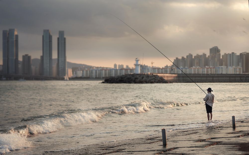 Fisherman standing on seashore near city at sunset