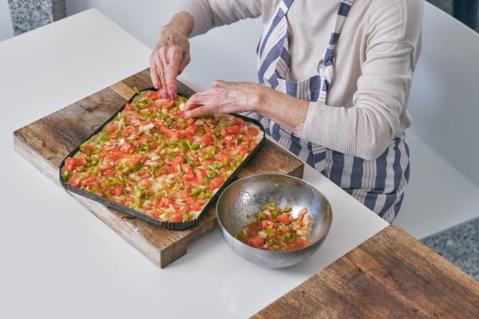 Crop aged woman adding vegetables to pizza
