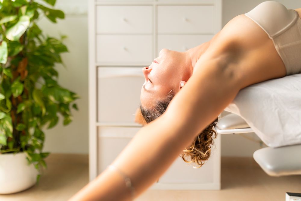 A woman performs a relaxing arm, back, and shoulder stretching exercise lying on a table at a physical therapy clinic