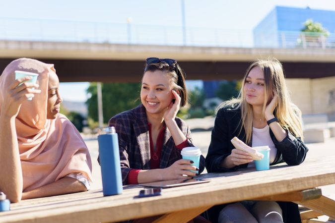 Smiling female friends sitting outside with warm drinks and sandwiches, copy space