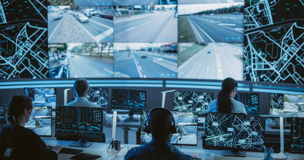 Dark Monitoring Room with Multiethnic Specialists Monitoring Surveillance Feeds and Coordinating Traffic Operations on Desktop Computers.