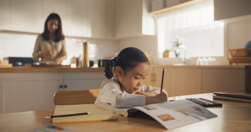 Portrait of a Korean Female Child Sitting at a Kitchen Table and Drawing While her Mother is Preparing Breakfast.