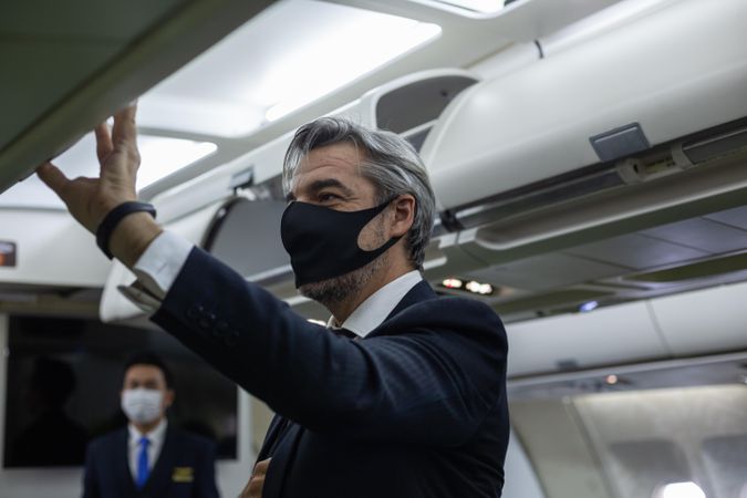 Grey haired male in mask putting away small suitcase in flight
