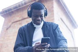Smiling Black man outside, wearing headphones and checking his phone 5kRQwG
