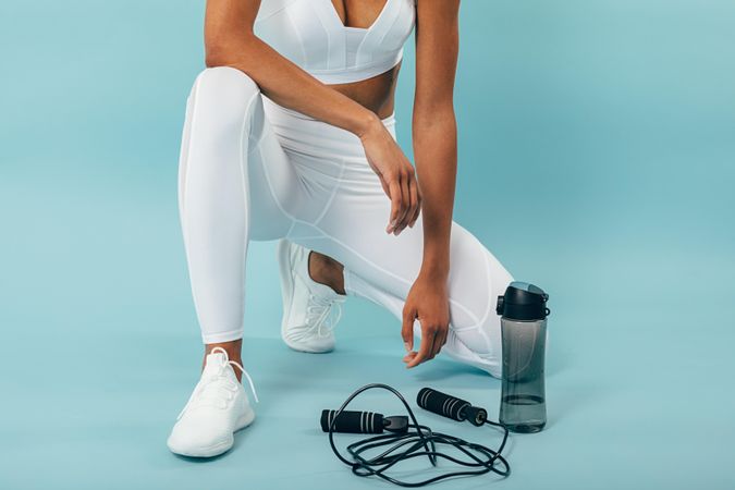 Fit woman kneeling down with water bottle and jump rope on blue background