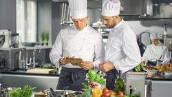 Two Famous Chefs Exchange Ideas About Video Blog Recipe they Watching on Tablet. They are Working in Big Restaurant Kitchen.