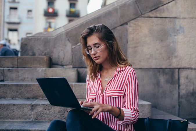 Serious young woman sitting on outdoor steps in European city working on her laptop