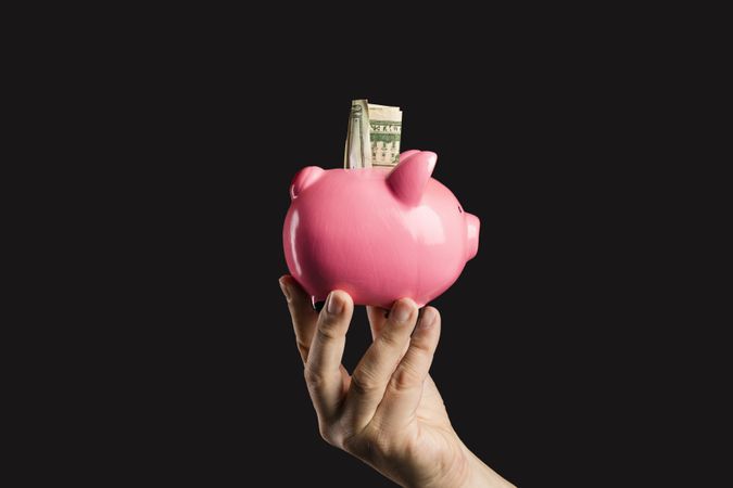 Hand holding a pink piggy bank with a dollar bill on a dark background.