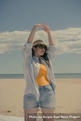 Woman in denim shorts wearing hat and sunglasses raising both arms and standing on beach 0WM6W4