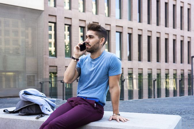 Man with beard in blue t-shirt speaking on phone while sitting outside building